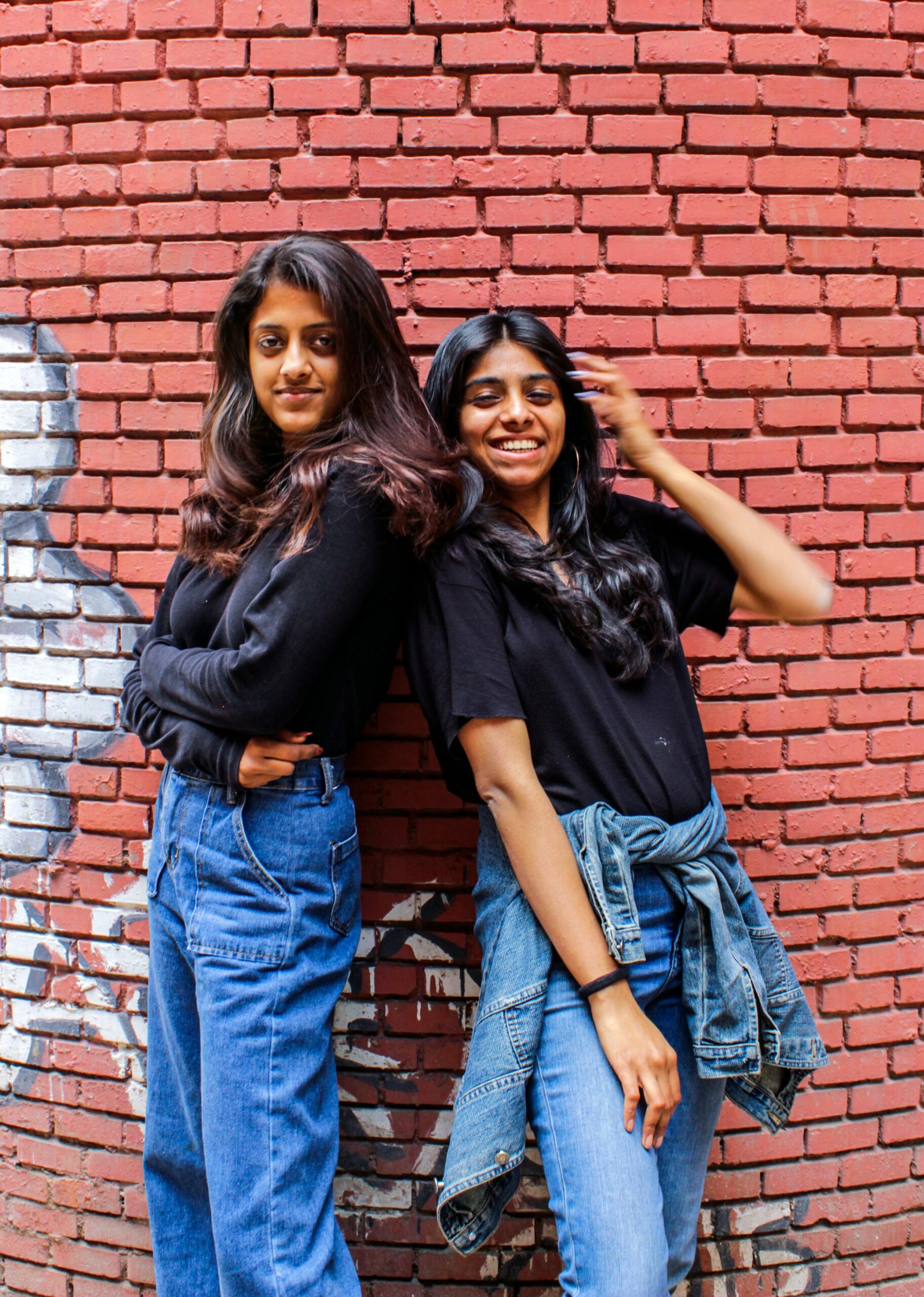 Two women in casual outfits posing confidently against a graffiti brick wall.